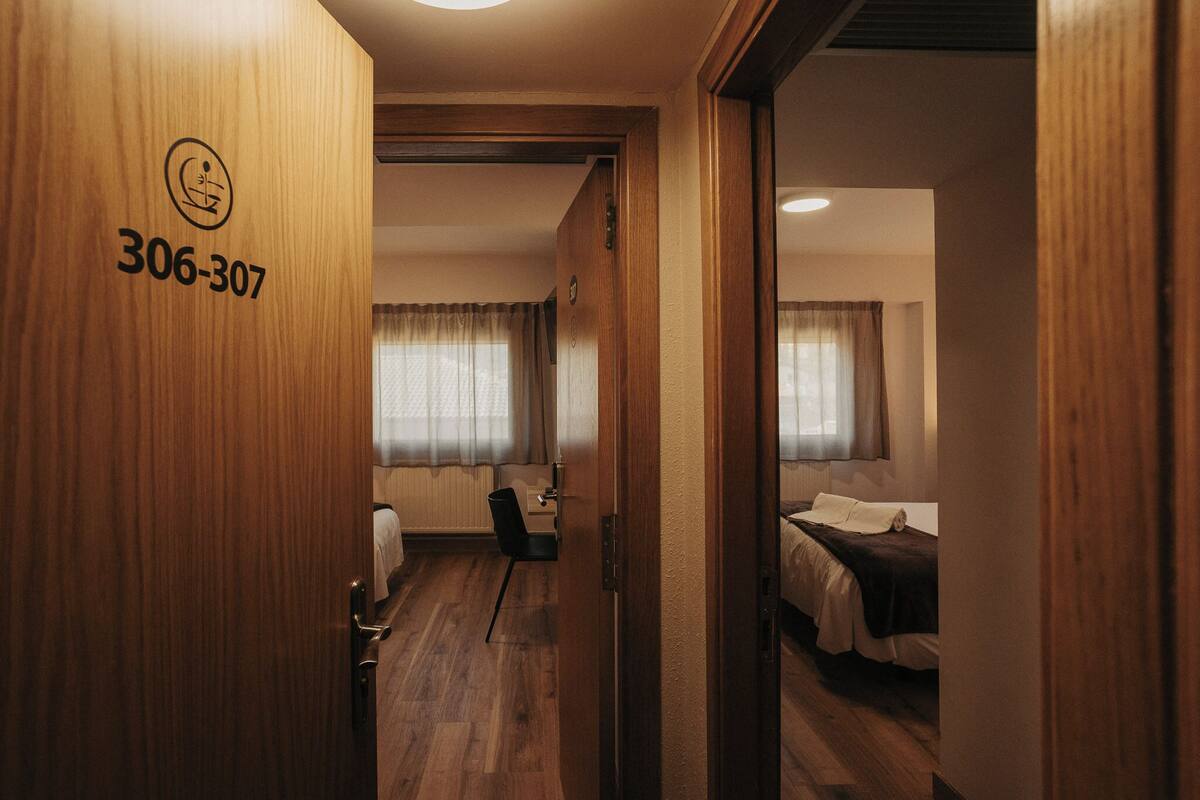 A hotel room with a wooden door marked '306-307,' a bed, a chair, a desk, and a window with curtains.