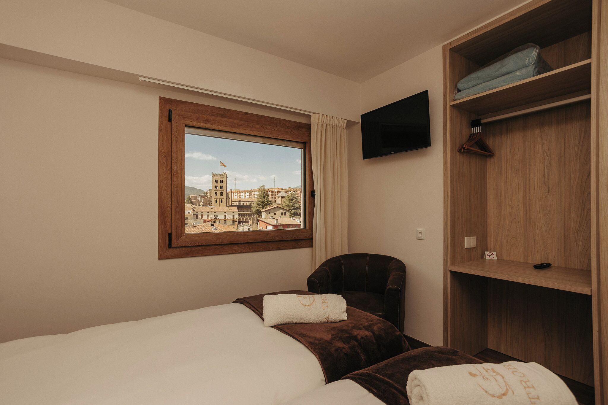 Habitación de hotel con cama, una silla, televisor montado en la pared y ventana con vista a edificios.