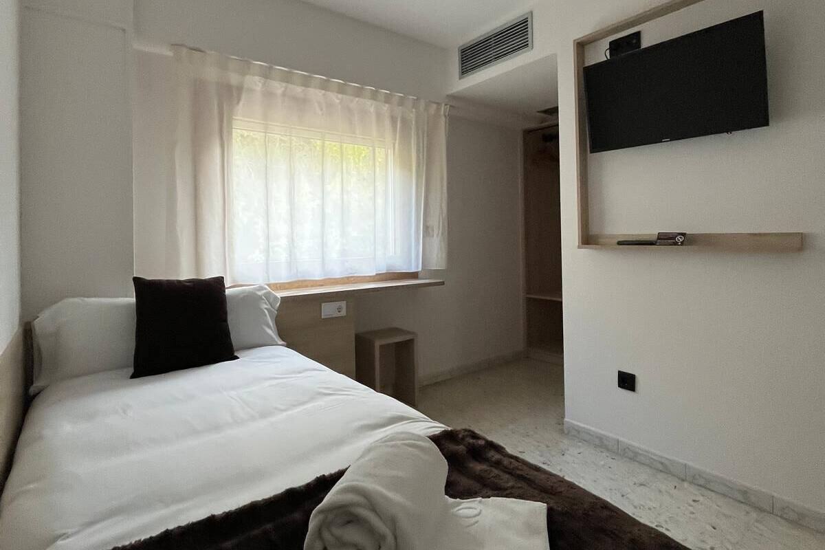 A hotel room with a bed, a TV mounted on the wall, a window with curtains, and a small bedside table.