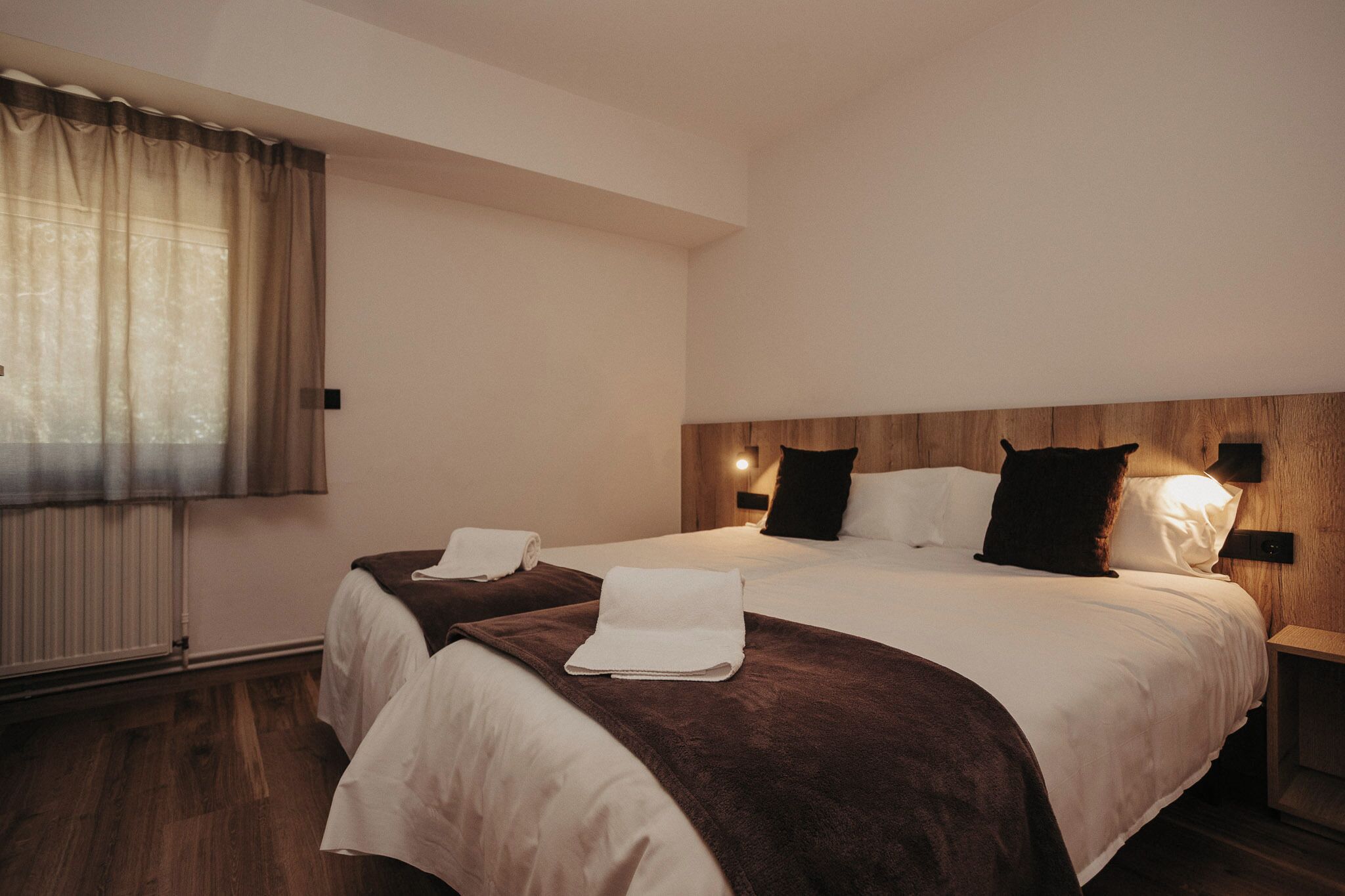 Habitación de hotel con una cama grande, dos mesitas de noche, una ventana con cortinas y un cabecero de madera.