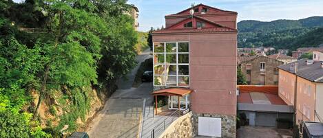 Un edificio moderno con fachada de vidrio, techo rojo y base de piedra. Se ubica en una zona ondulada con vegetación y otros edificios al fondo.