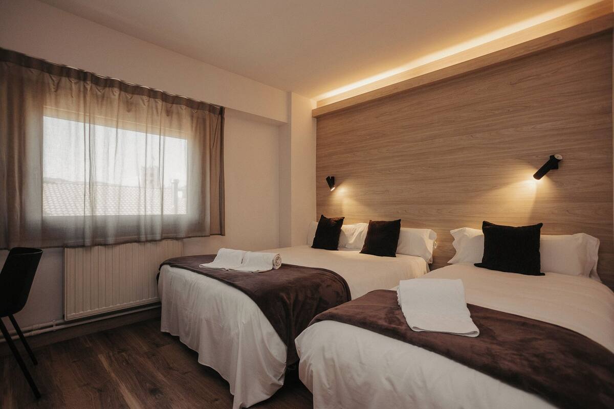 A hotel room with two beds, a window with sheer curtains, a wooden headboard, and a wall-mounted light fixture.