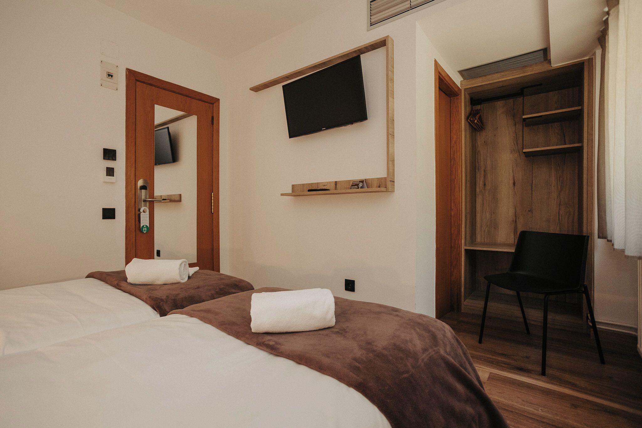 Habitación de hotel con dos camas, un televisor montado en la pared, un armario de madera y una silla.