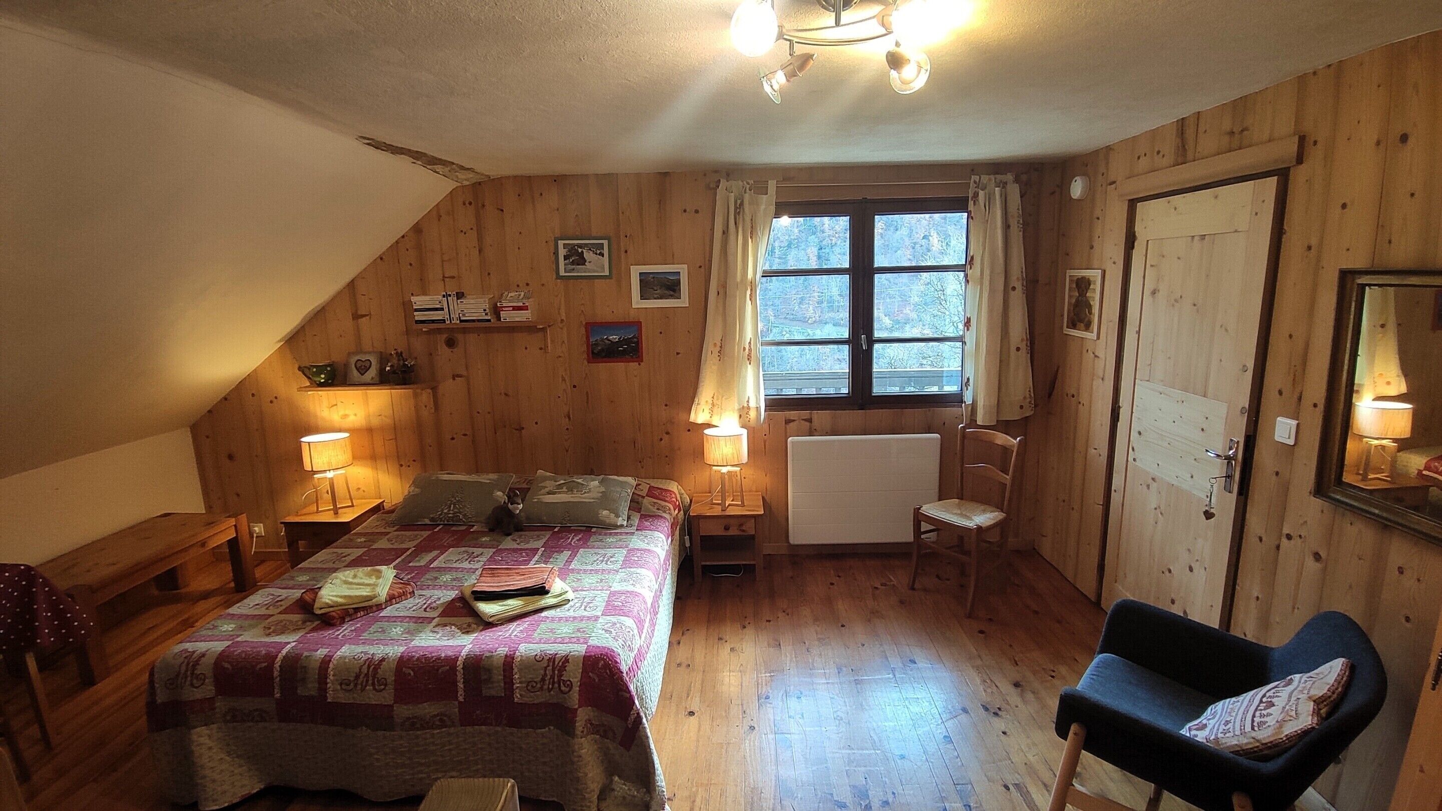 A cozy attic bedroom with a sloped ceiling, wooden walls, a bed with a quilt, two bedside lamps, a chair, and a window with curtains.