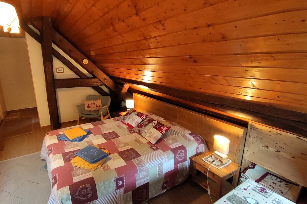 Un dormitorio acogedor en el ático con techo de madera, una cama con colcha, una mesita de noche y una silla.