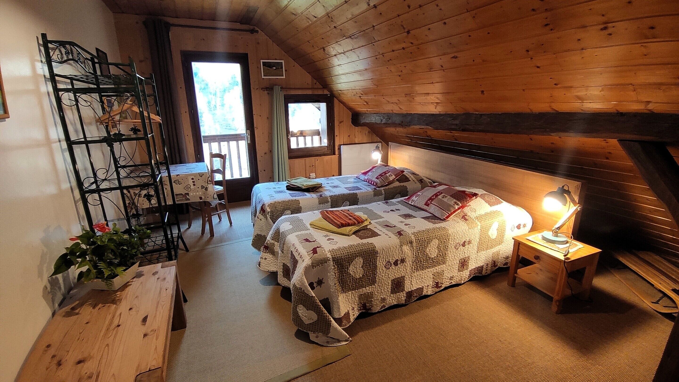 A cozy attic bedroom with two beds, a wooden desk, a chair, and a window.