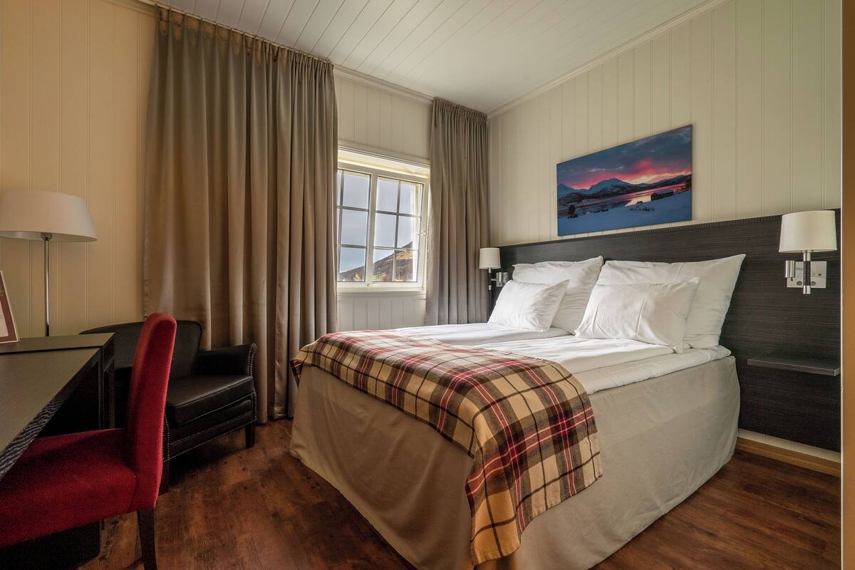 Ein Hotelzimmer mit Bett, Schreibtisch, Stuhl, einem Fenster mit Vorhängen und einem Bild an der Wand.
