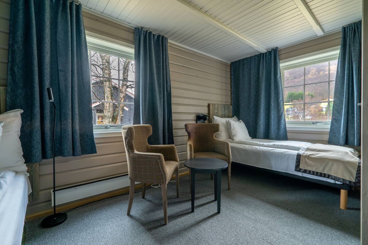 Ein Zimmer mit einem Bett, zwei Stühlen, einem kleinen Tisch und großen Fenstern mit Vorhängen.