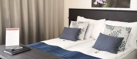 A hotel room with a bed, a desk, and a chair. There is a sign on the desk and a painting on the wall.