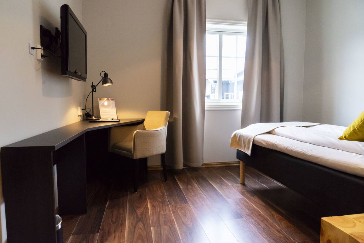 Ein Schlafzimmer mit einem Bett, einem Schreibtisch mit Stuhl, einer Lampe und einem Fenster mit Vorhängen.