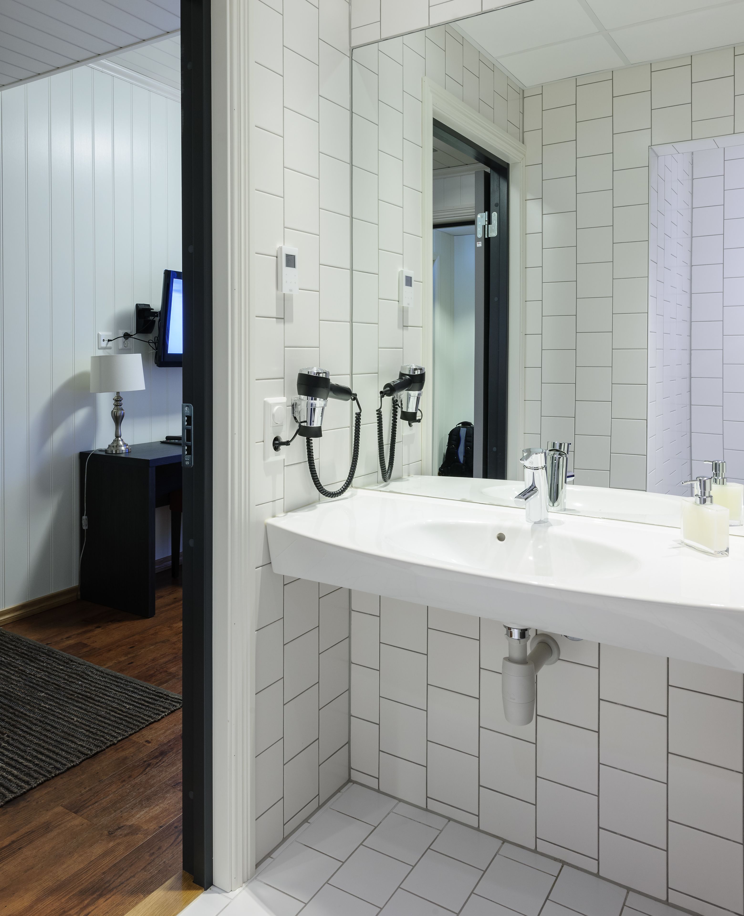 A modern bathroom with a large white sink, two hair dryers mounted on the wall, a mirror, and a small table with a lamp.