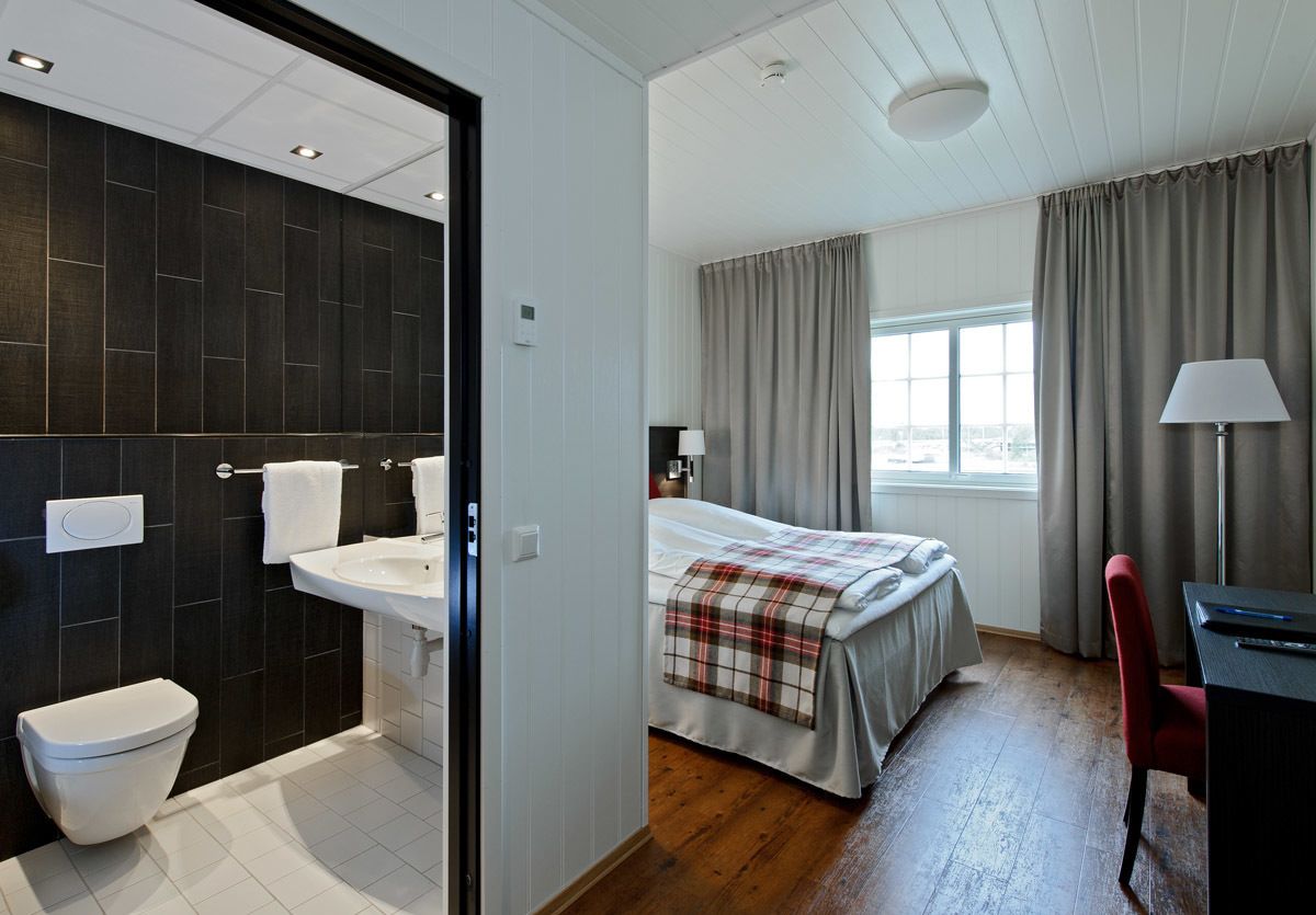 A modern hotel room with a dark tiled bathroom, a bed with a plaid blanket, a desk with a lamp, and a view of the city through large windows.