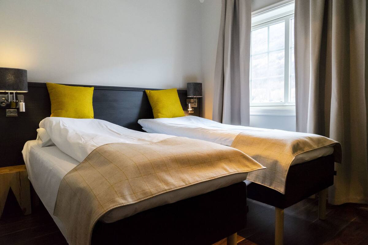Ein Doppelbett mit weißer Bettwäsche, ein schwarzer Kopfteil und zwei gelbe Kissen. Zwei Nachttischlampen und ein Fenster mit Vorhängen.