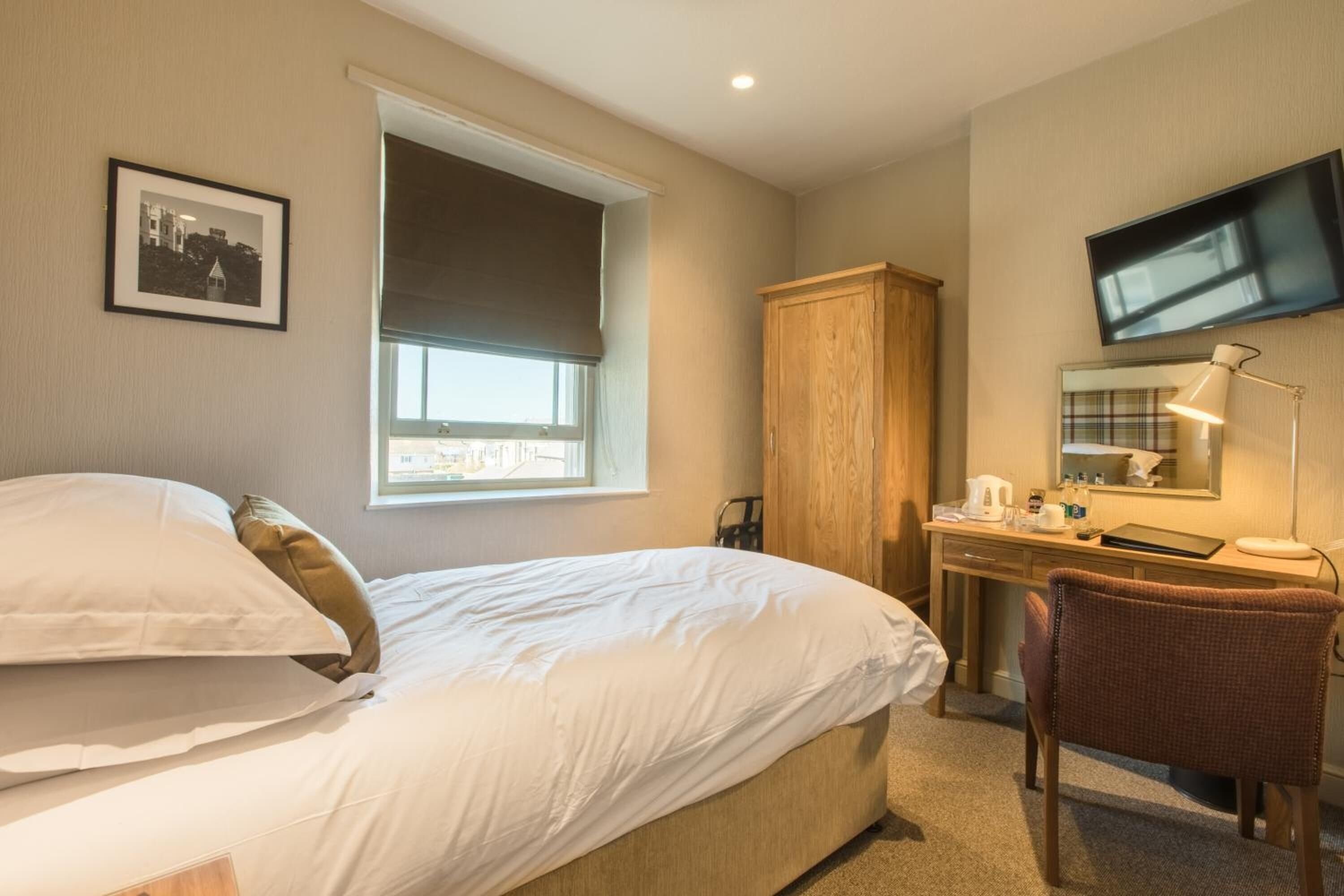 A hotel room with a bed, a desk, a chair, a TV, a window with a blind, and a framed picture on the wall.