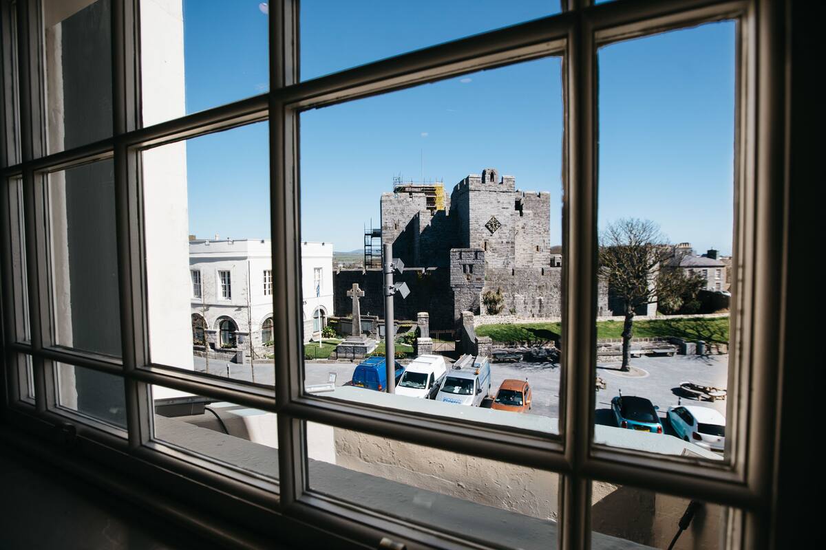 Blick durch ein Fenster auf ein schlösserartiges Gebäude mit einem Parkplatz davor.