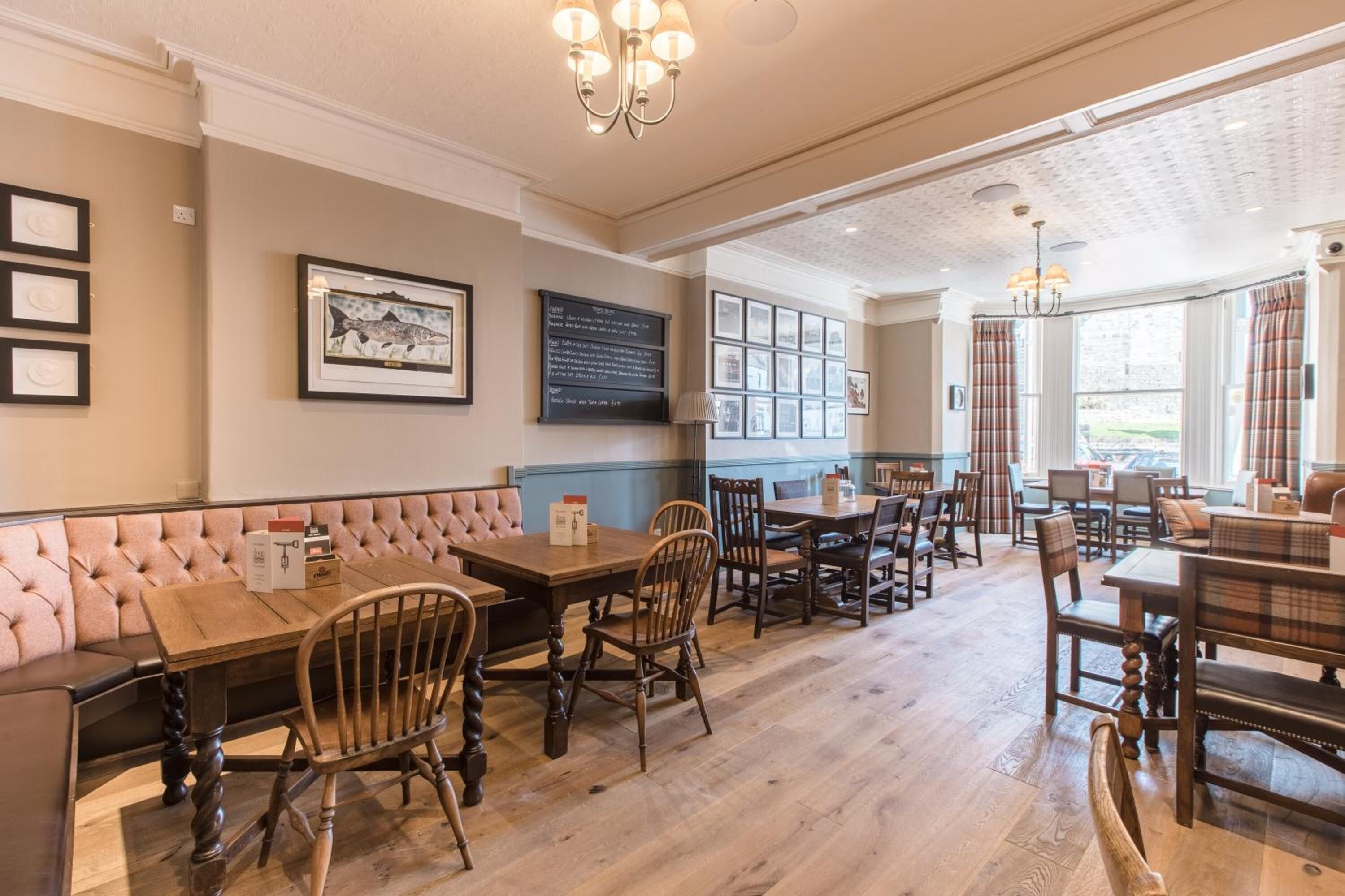 A restaurant with wooden tables and chairs, a cushioned bench, framed pictures on the walls, and a chandelier.