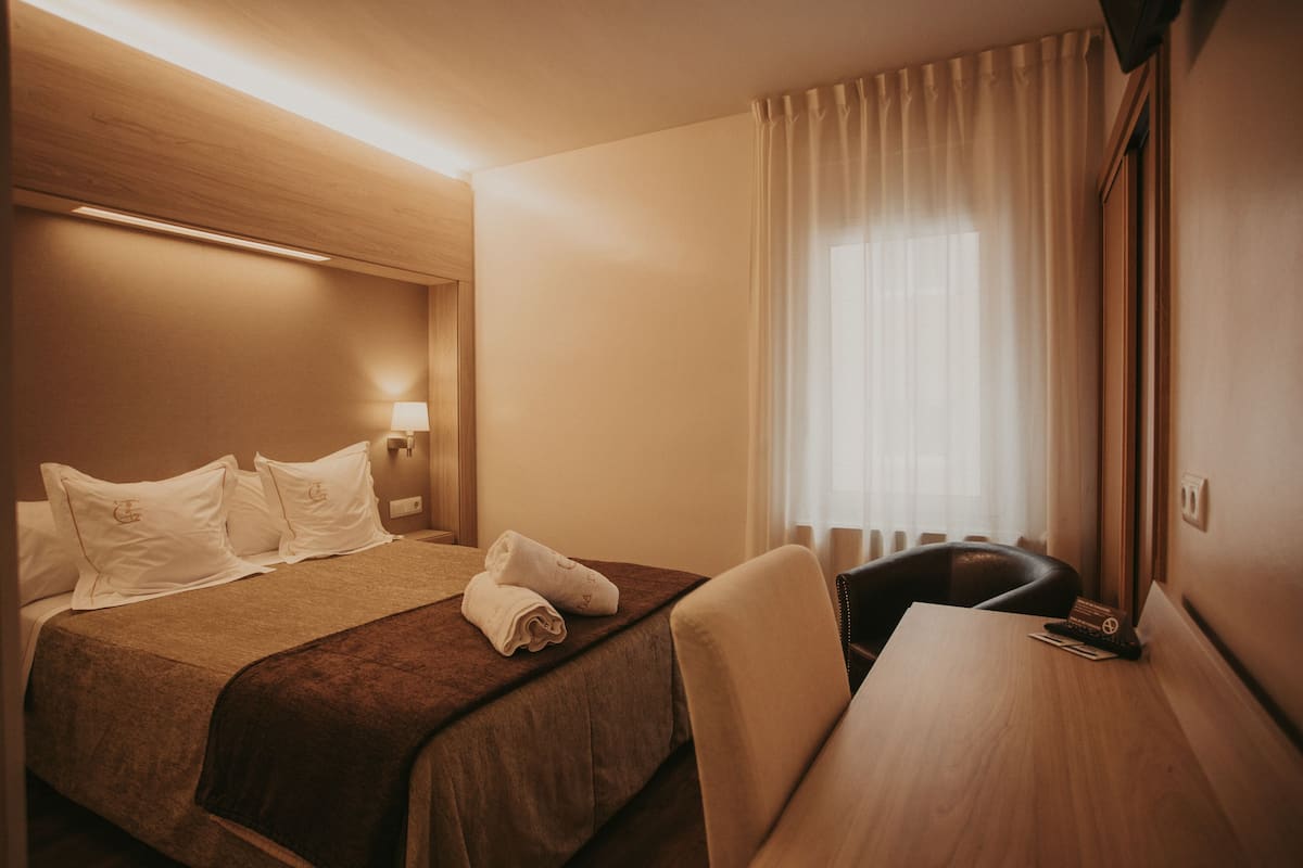 A hotel room with a bed, a desk, a chair, a lamp, and a window with curtains.