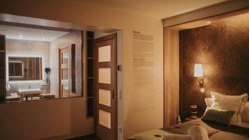 A hotel room with a large bed, a bedside table with a lamp, a wall-mounted information panel, and a view into the bathroom.