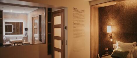A hotel room with a large bed, a bedside table with a lamp, a wall-mounted information panel, and a view into the bathroom.