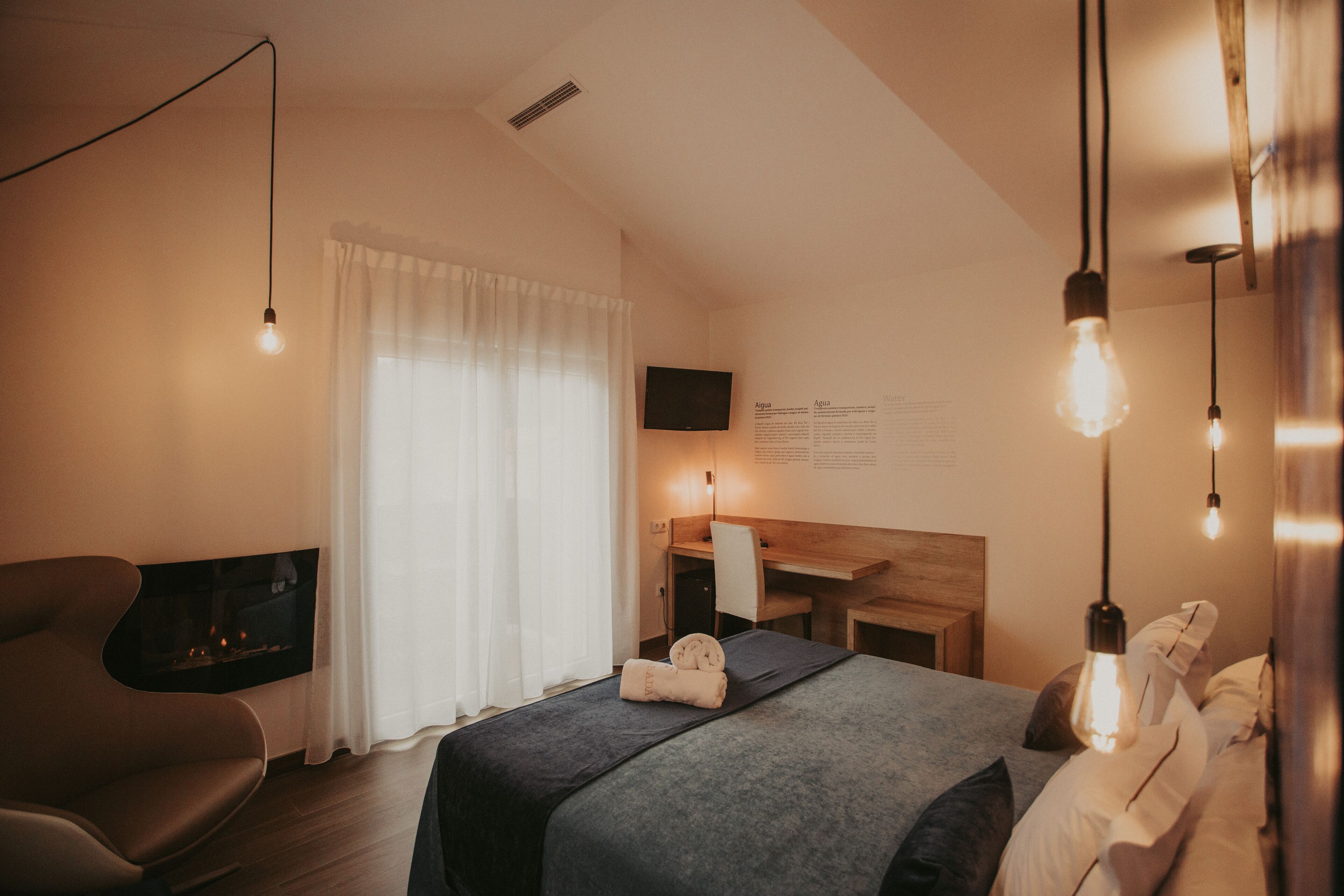 A modern hotel room with a bed, a desk, a chair, a flat-screen TV, and a fireplace.