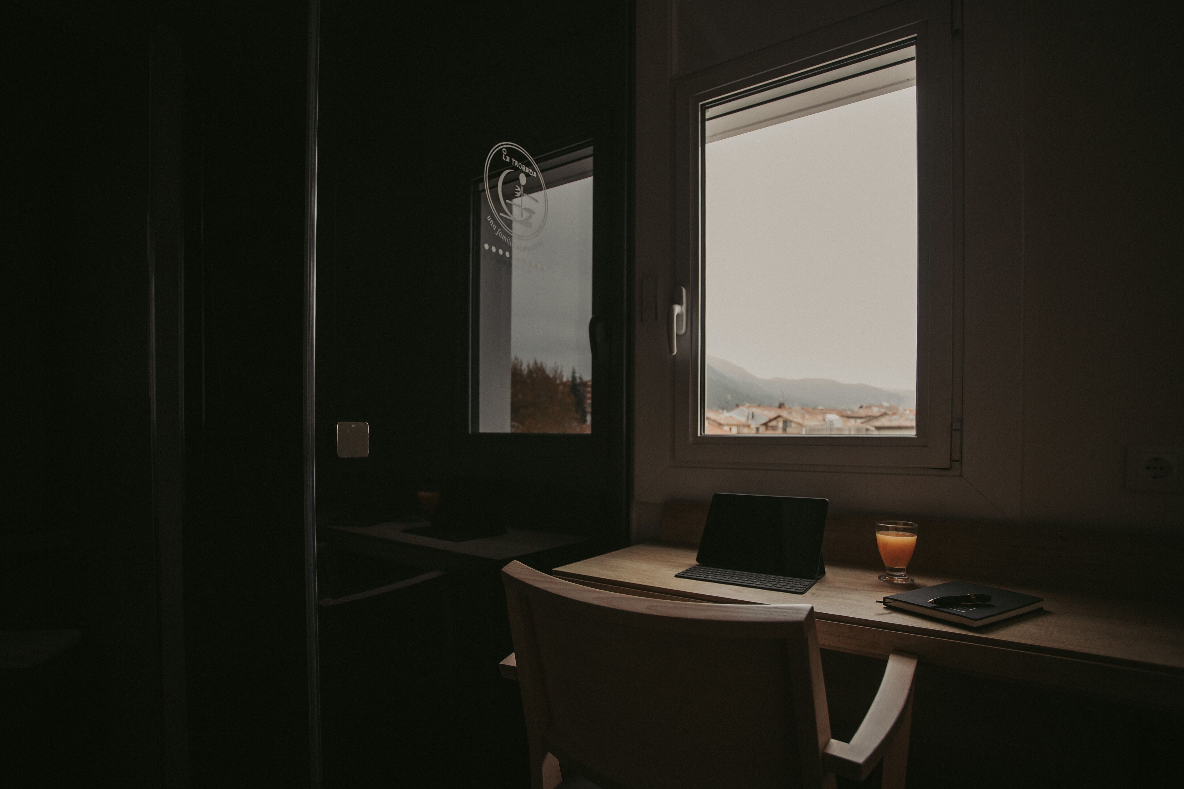 Una habitación tenuemente iluminada con un ordenador portátil sobre un escritorio, un vaso de zumo de naranja y vistas a las montañas a través de una ventana.