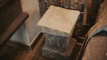 A stone bench with a rough texture, positioned in a corner with a wooden floor and a stone wall in the background.