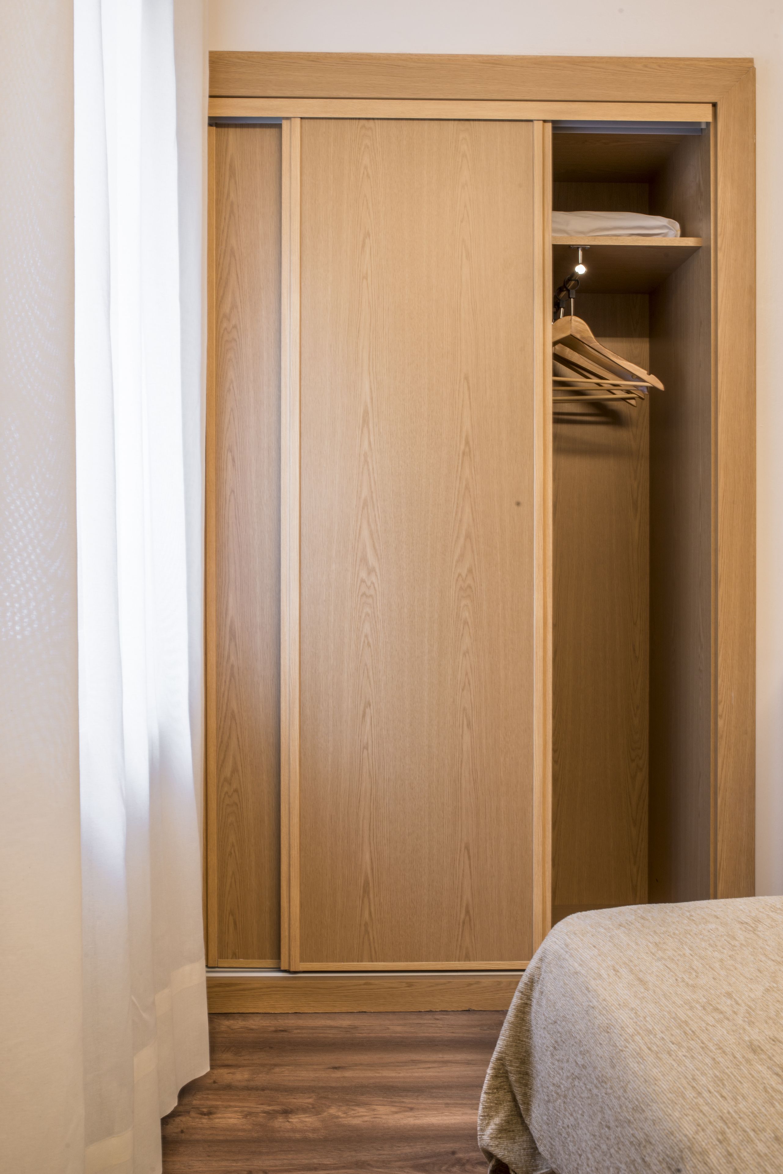 Un armario de madera con espacio para colgar, una cama con manta color beige y suelo de madera.