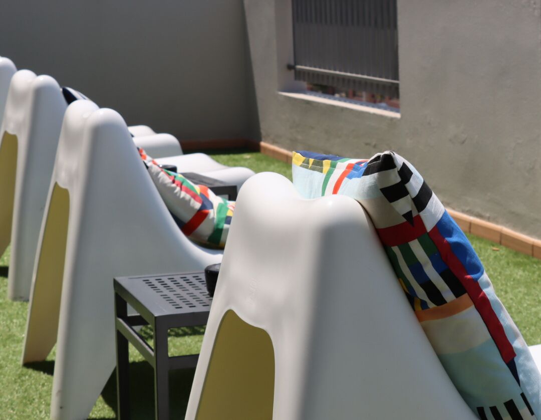 Una fila de sillas de plástico blancas con mantas de colores, dispuestas al aire libre frente a una pared y una ventana.