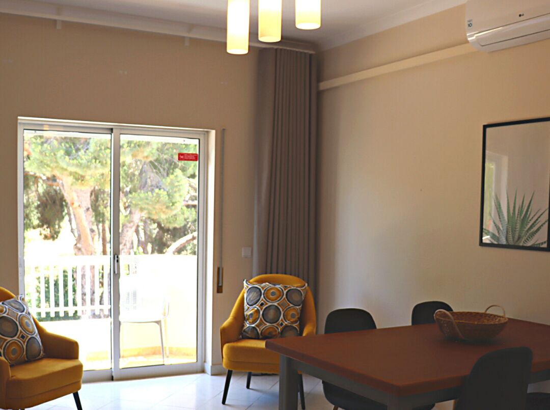 Een moderne kamer met een eettafel, gele fauteuils en een groot raam met uitzicht op bomen en een witte schutting.