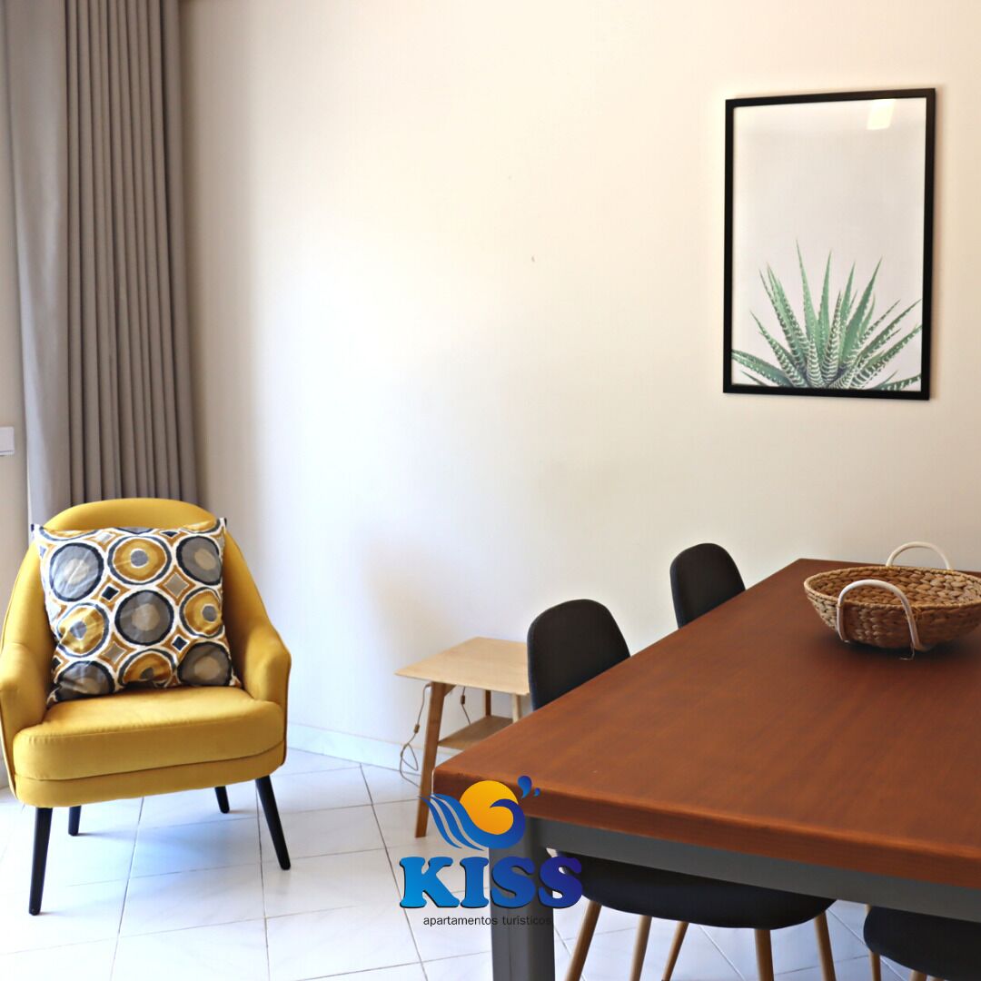 A dining area with a wooden table, black chairs, a yellow armchair with patterned cushions, a woven basket, and a framed plant on the wall.
