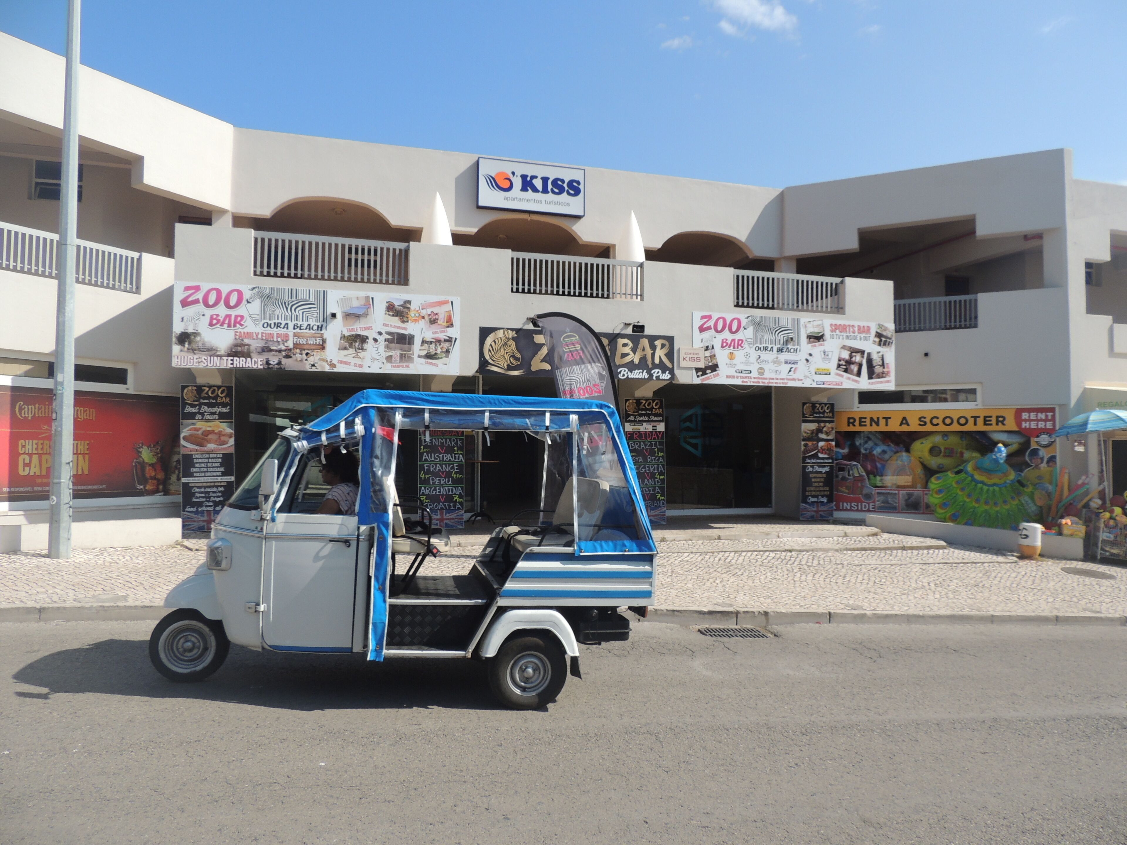 A tuk-tuk parked in front of a building with multiple signs, including 'KISS' and 'ZOO BAR'.