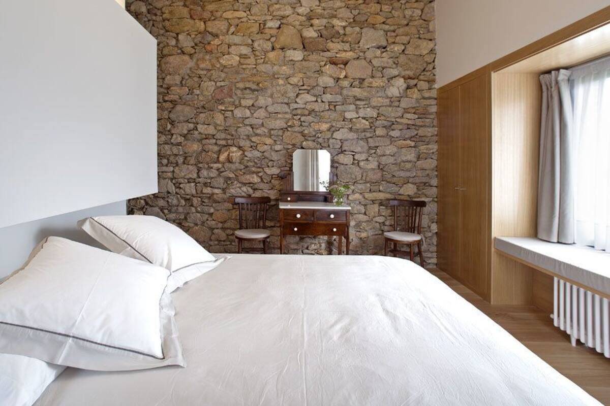 A bedroom with a stone wall, a wooden dresser, a mirror, and a window with curtains.