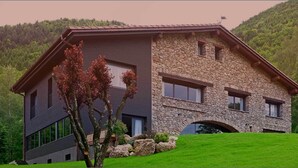 A modern two-story house with a stone facade and a sloped roof, surrounded by lush greenery and hills. - L'Hotelet del Bac (Camprodon)