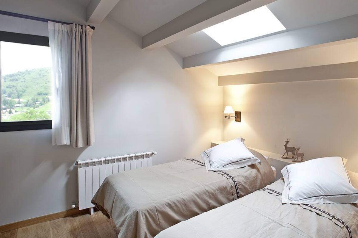 A bedroom with two beds, a desk, and a window with a view of greenery.