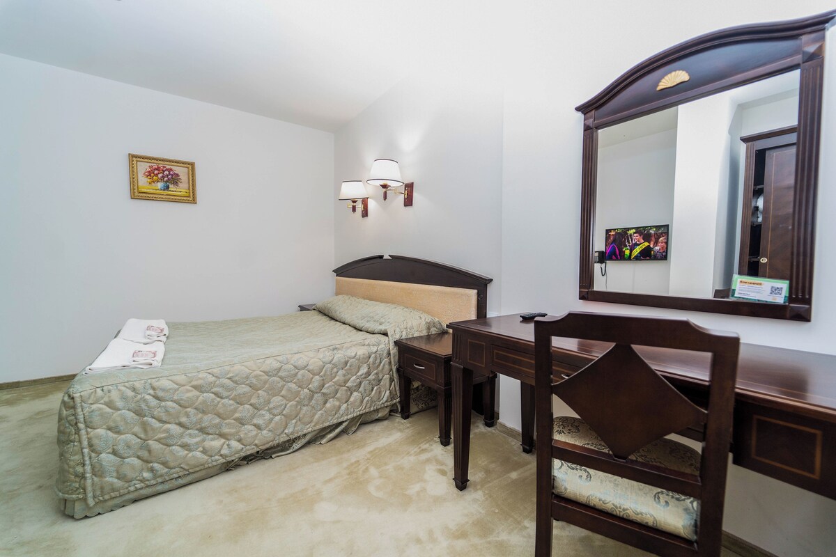 A bedroom with a bed, a desk, a chair, a mirror, and a wall-mounted lamp.