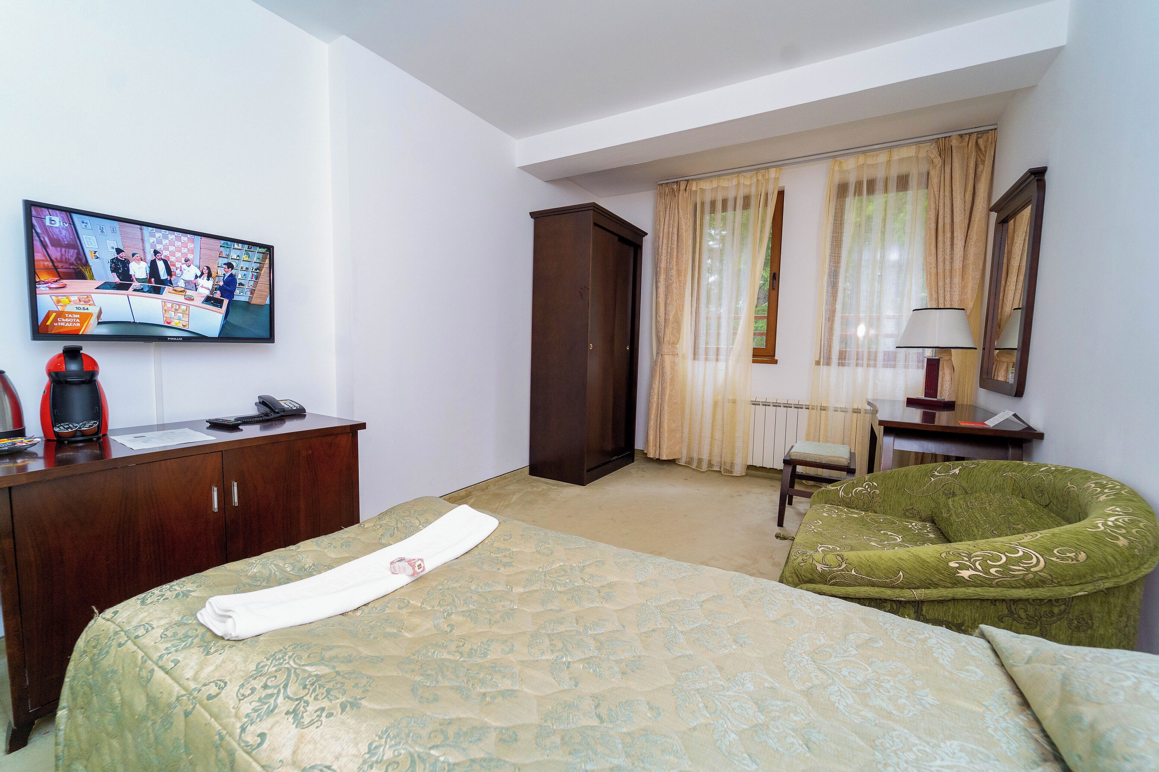 A hotel room with a bed, a desk with a television, a green armchair, and a wardrobe.