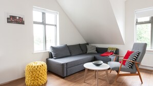 A modern living room with a grey sofa, a yellow ottoman, a wooden chair, and a small white table. - Mengi Apartments (Reykjavik)