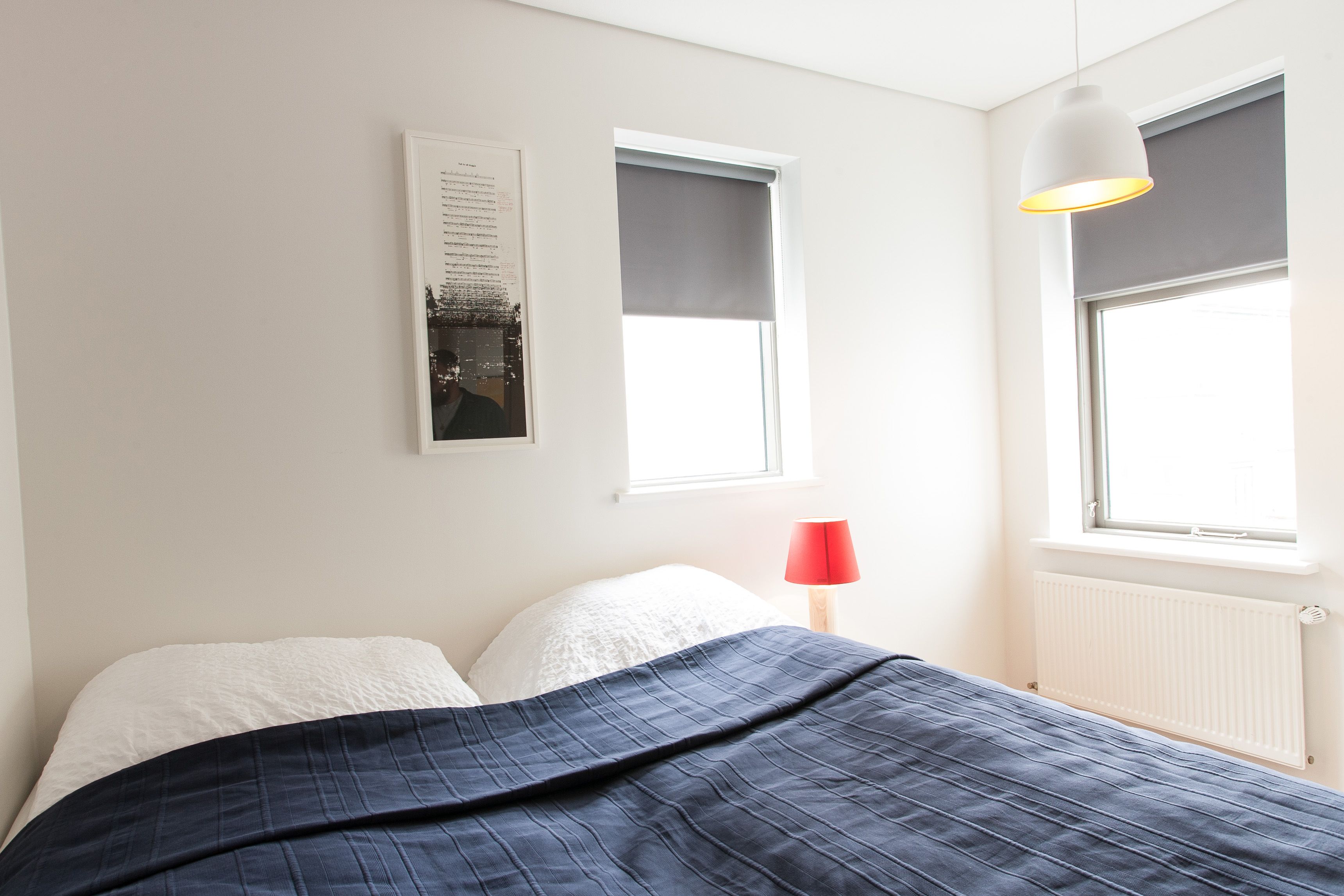 Een slaapkamer met een bed, een wandlamp, een raam met zonwering en een radiator.