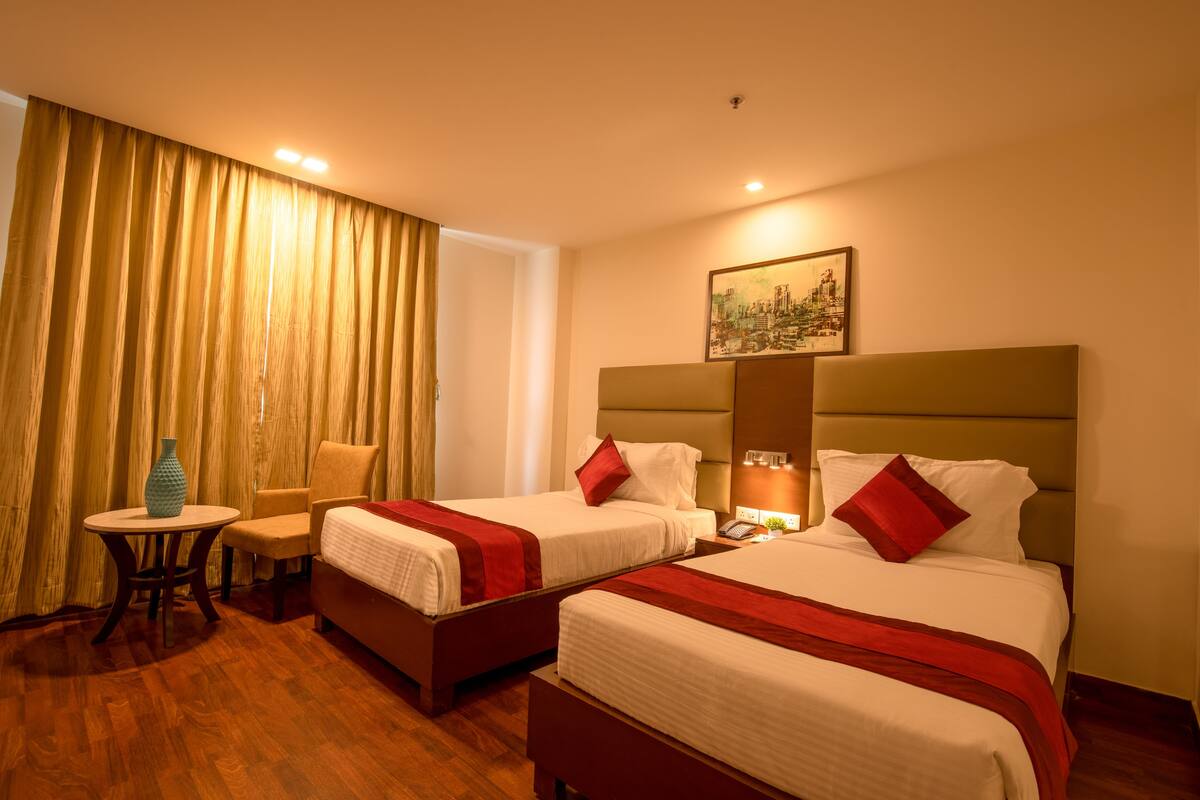 A hotel room with two beds, a small table, a chair, and a painting on the wall.
