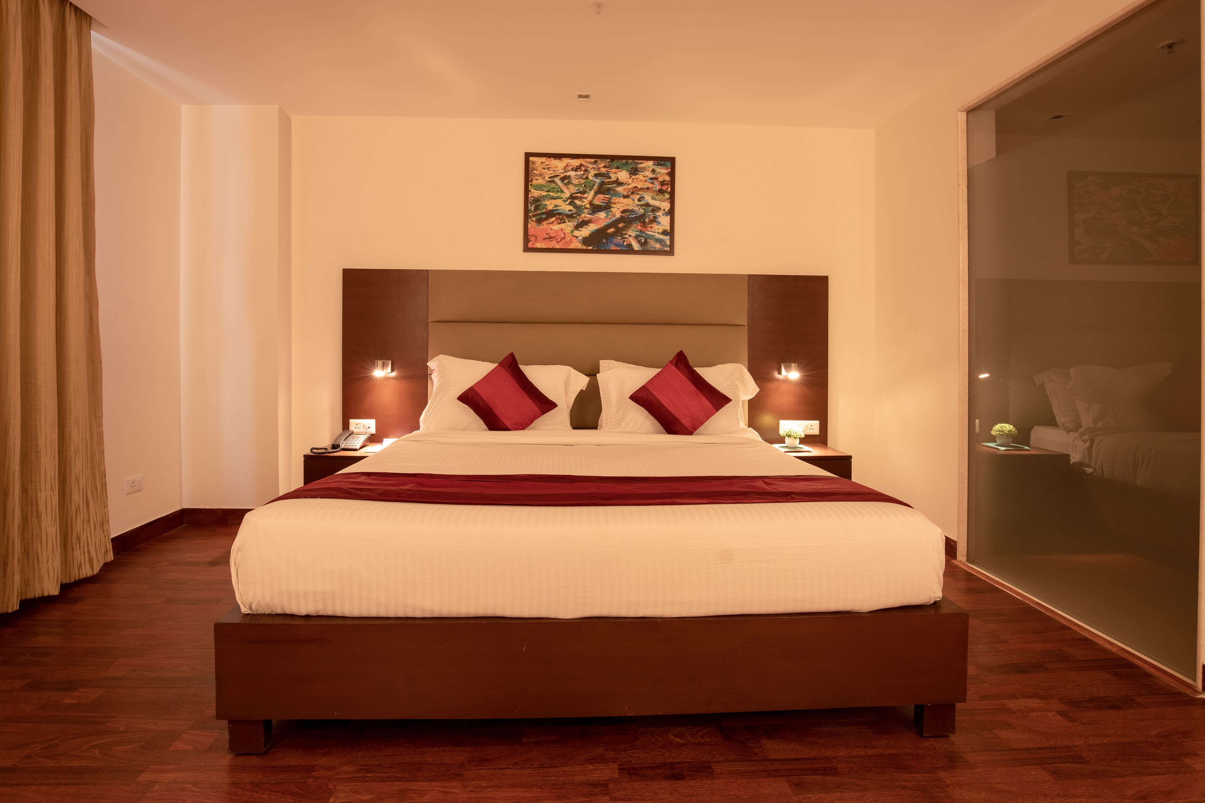 A large bed with a wooden frame, white bedding, and red pillows. There are two bedside tables with lamps. A painting is on the wall above the bed.
