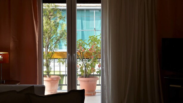 A room with a sofa, a lamp, a TV, and a sliding glass door leading to a balcony with potted plants. - Shiny Modern Apartment • 1' to Metro Station (Athens)