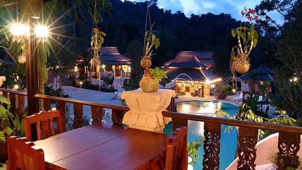 A wooden table and chairs on a balcony overlooking a pool, with hanging plants and a traditional building in the background. - Grand Orchid Resort (Ko Chang)
