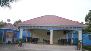 Un área de estar al aire libre cubierta, con suelo de baldosas, rodeada de sillas y mesas, y un edificio con techo rojo y paredes azules.
