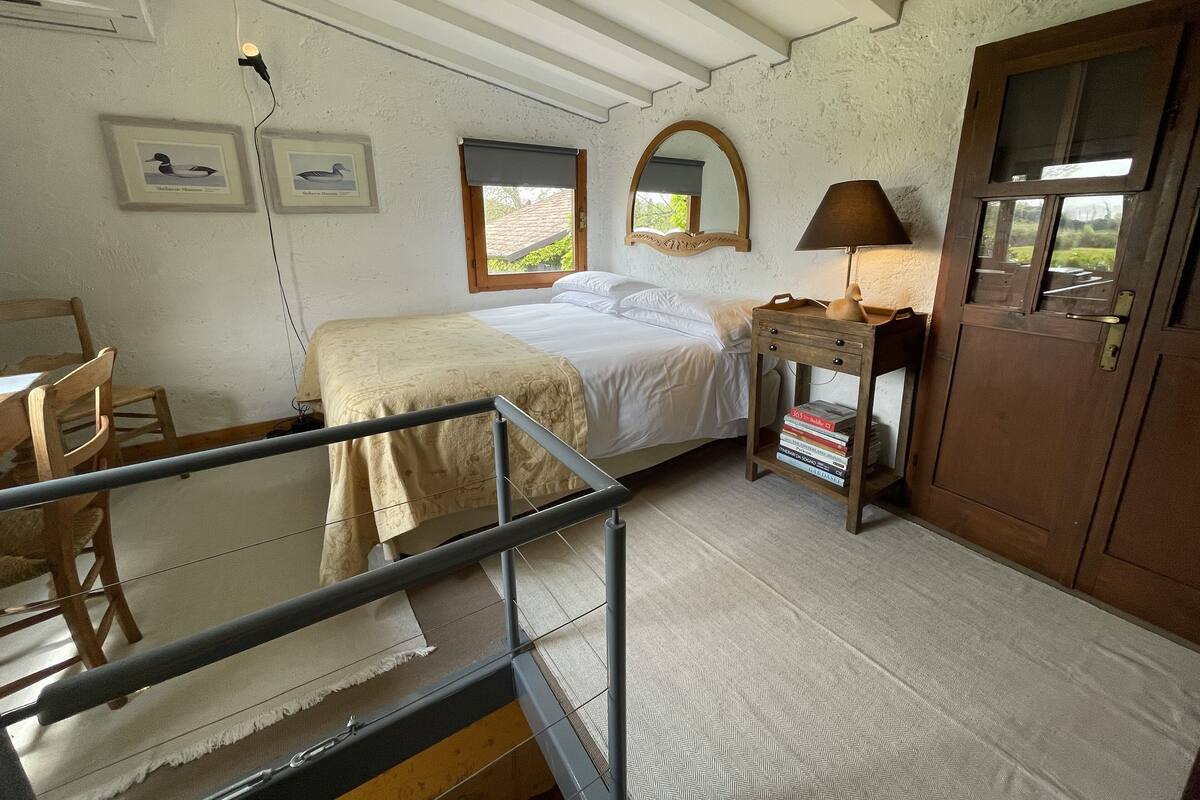 Une chambre à coucher avec un lit, une table de chevet avec des livres, une lampe, une chaise en bois, un miroir et une fenêtre donnant sur un paysage verdoyant.