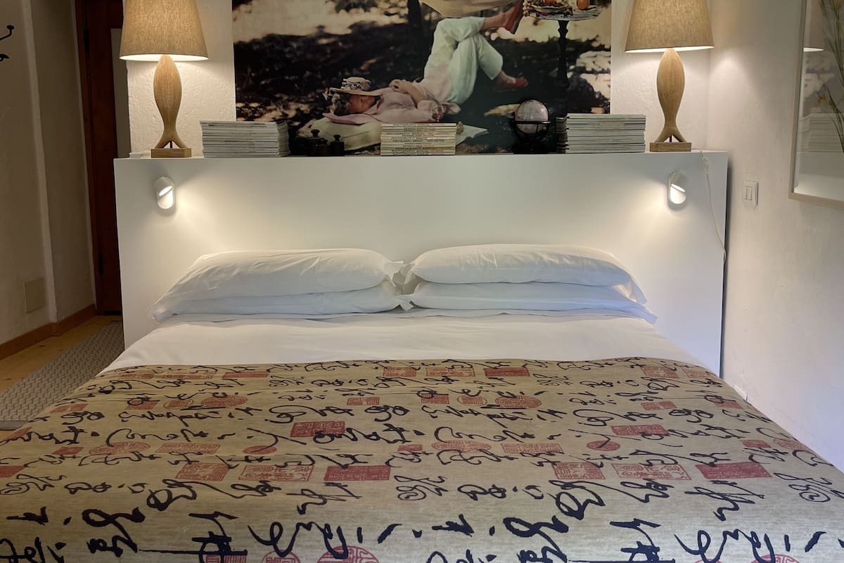 Une chambre à coucher avec un lit, deux lampes de chevet, une étagère fixée au mur avec des livres, et un tableau encadré au-dessus du lit.