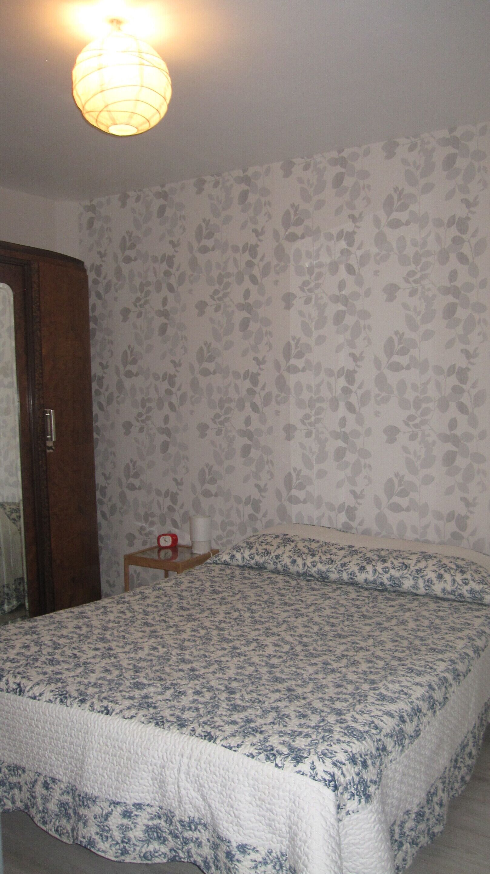 Ein Schlafzimmer mit einem Bett, einer Holztür, einem kleinen Tisch mit einer Uhr und floralem Wandpapier.