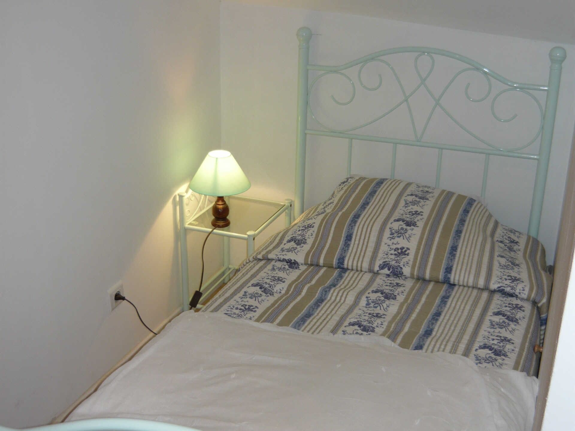 A small, single-bed room with a metal headboard, a bedside table with a lamp, and a floral-patterned bedspread.