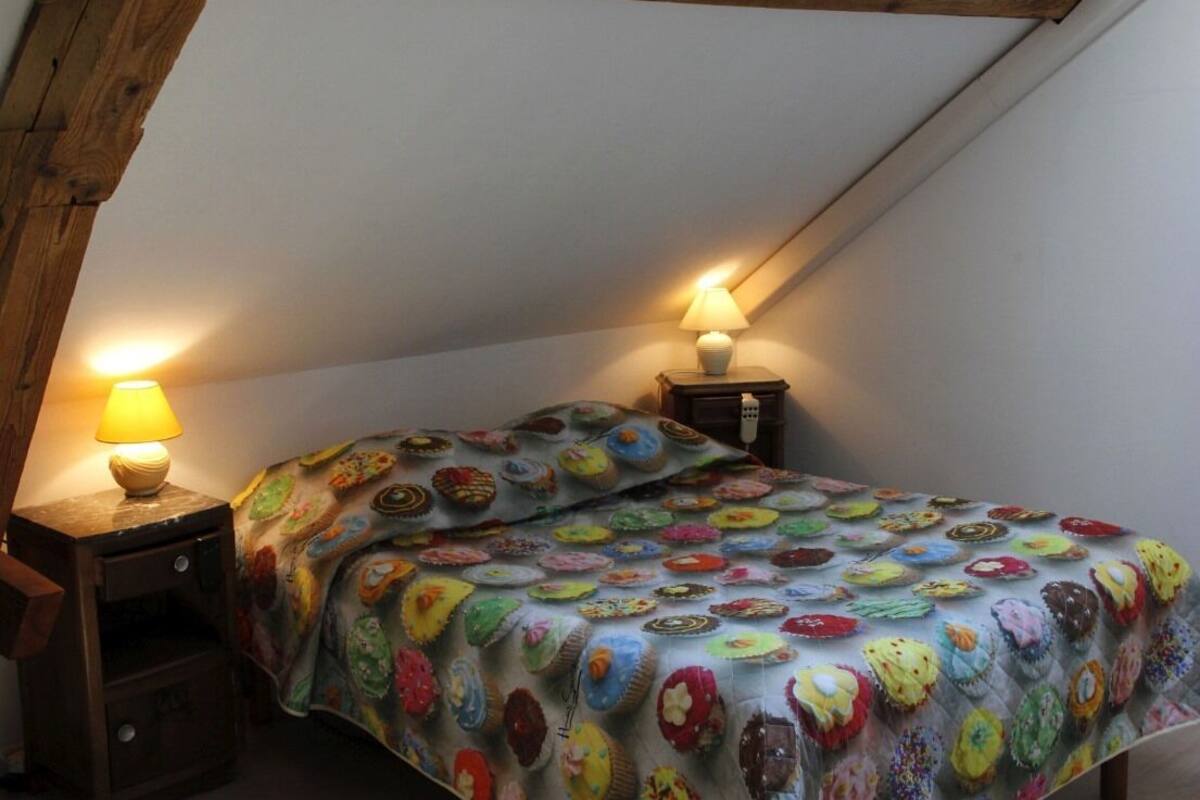 Une chambre avec un plafond en pente, un lit recouvert d’une couette colorée, deux lampes de chevet et une petite table de nuit en bois.