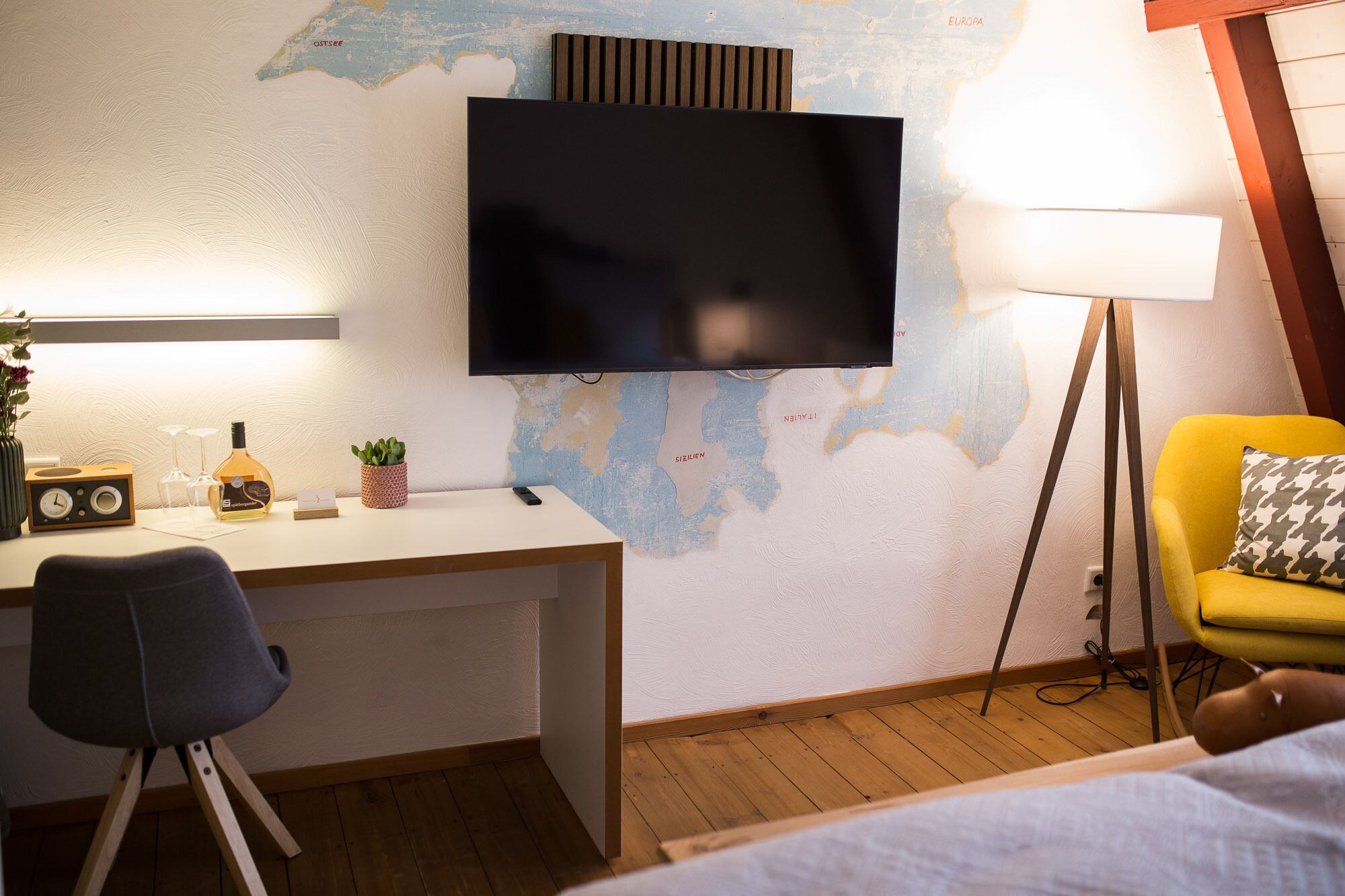 A hotel room with a wooden desk, a flat-screen TV mounted on the wall, a map, a lamp, and a yellow armchair.