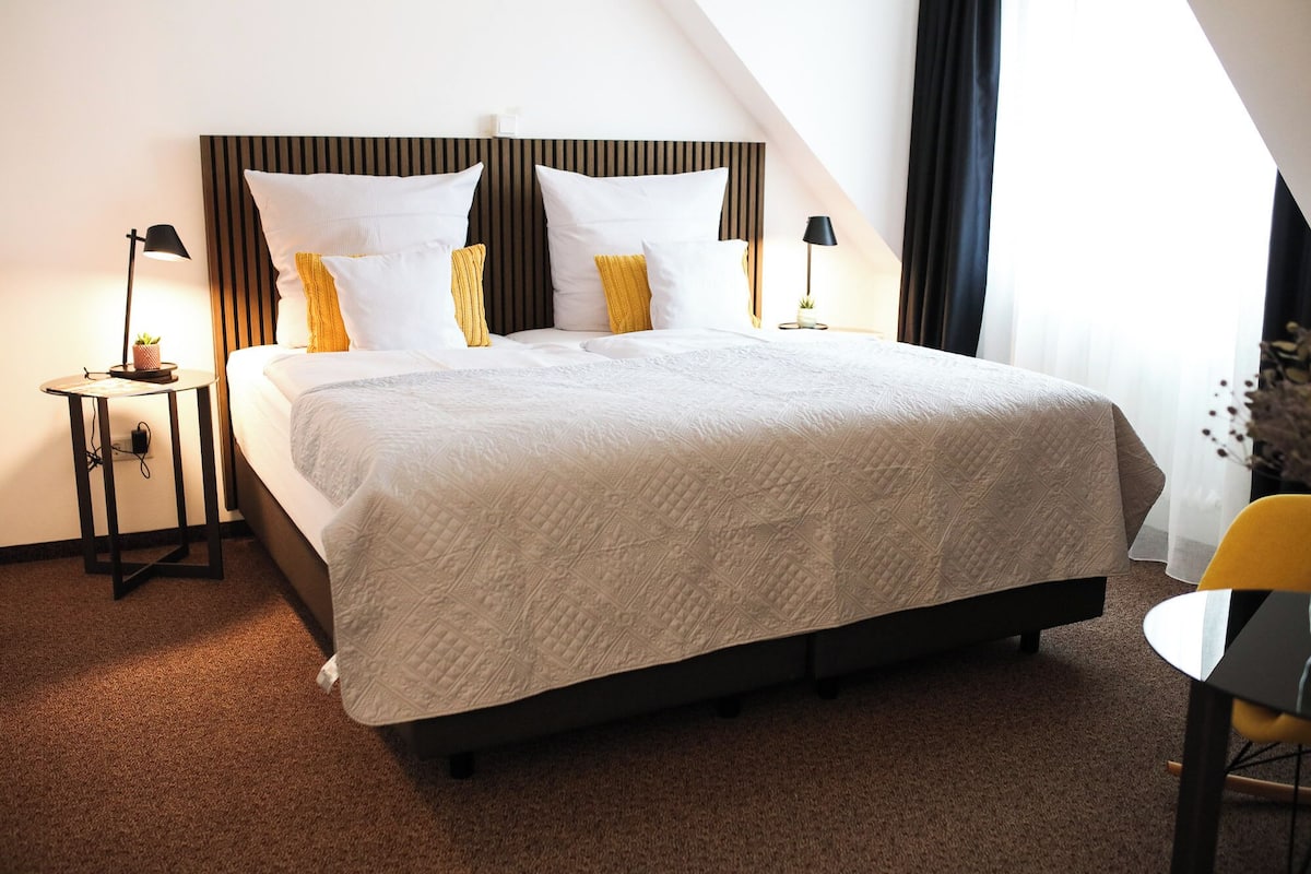 Een netjes ingerichte hotelkamer met een groot bed, twee nachtkastjes met lampen, een klein tafeltje met een stoel, en een raam met gordijnen.